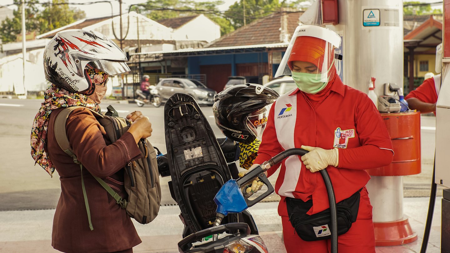 Gas station attendant fills motorcycle fuel tank with customer nearby.